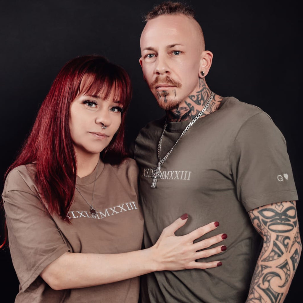 Tragebild von Partner T-Shirts, man sieht eine rothaarige mit einem braunem Shirt auf dem römische Zahlen in weiß drauf gestickt sind und daneben ein Mann der ein oliv farbenes Shirt trägt mit römischen Zahlen und am Arm hat er noch den Buchstaben G mit einem Herz